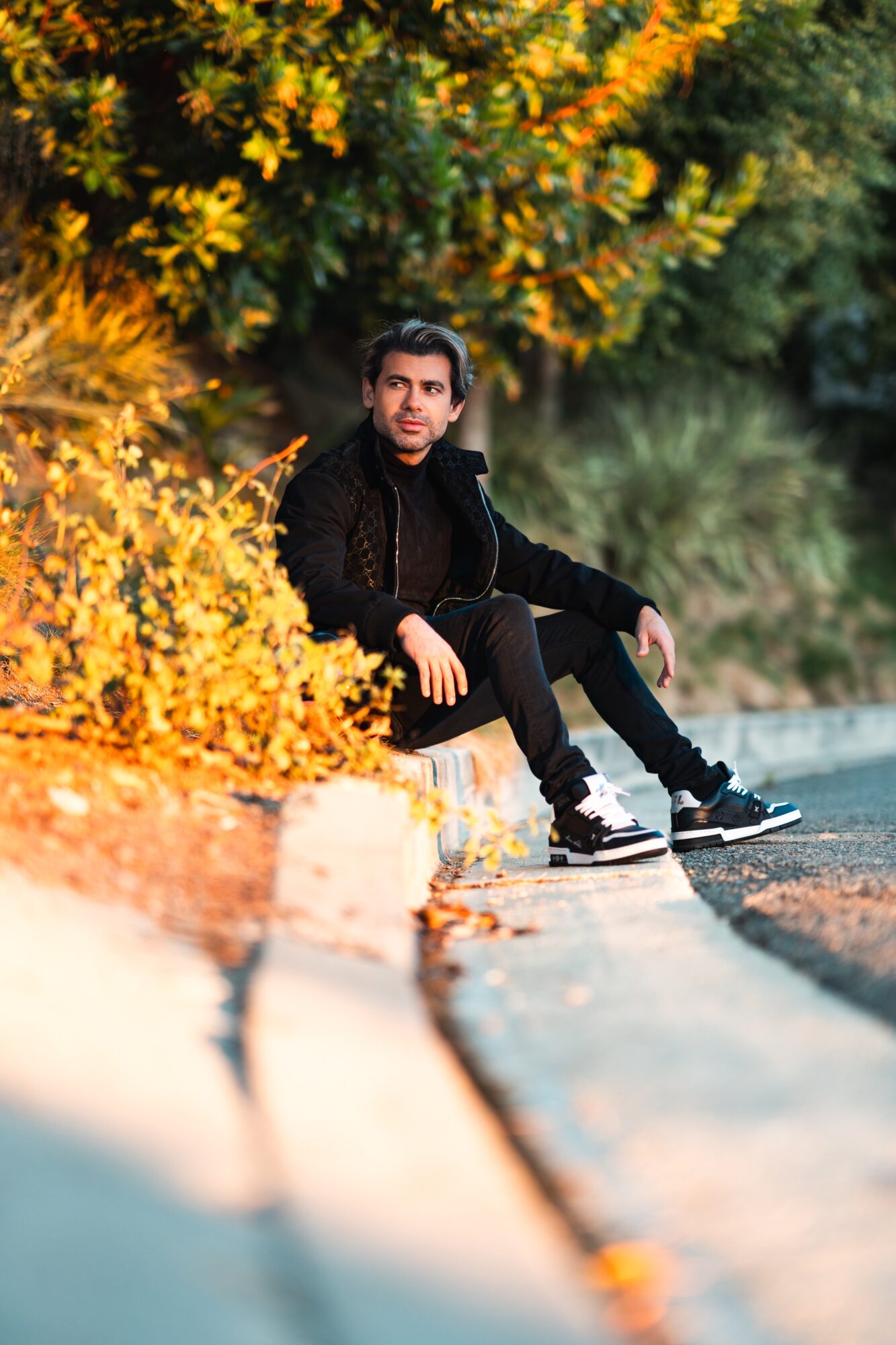 Young man sitting on a curb with yellow foliage behind him, wearing black clothing and sneakers, looking at the camera.