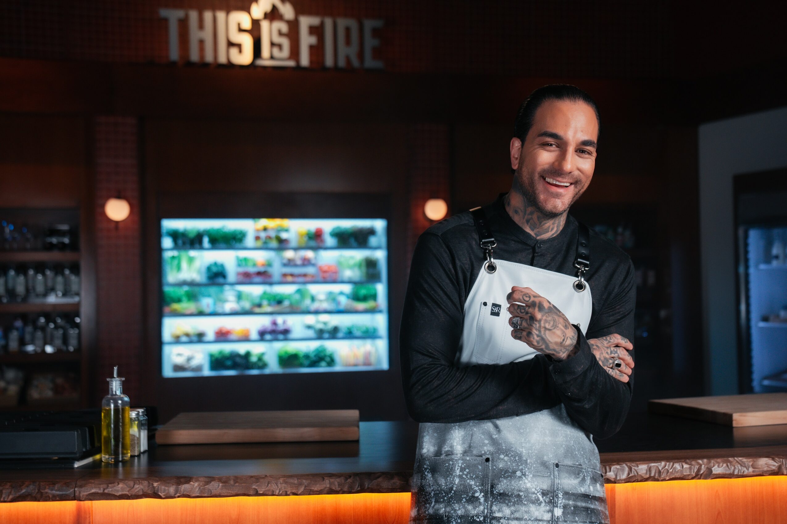 Smiling man with tattoos wearing an apron behind a bar counter in a restaurant or cafe setting.