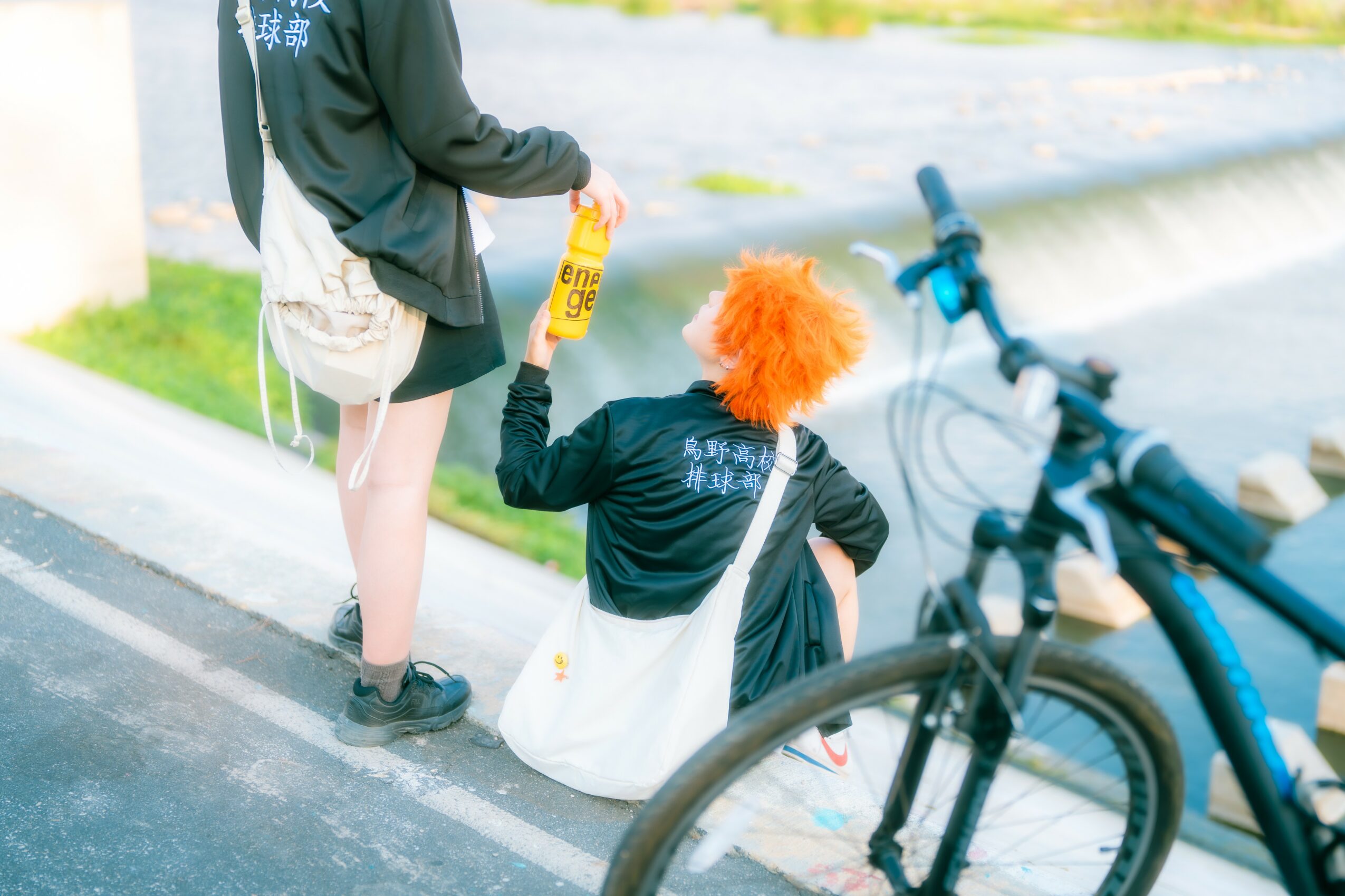Person with orange hair sitting on the ground near a bicycle, holding a yellow bottle, with another person standing nearby.