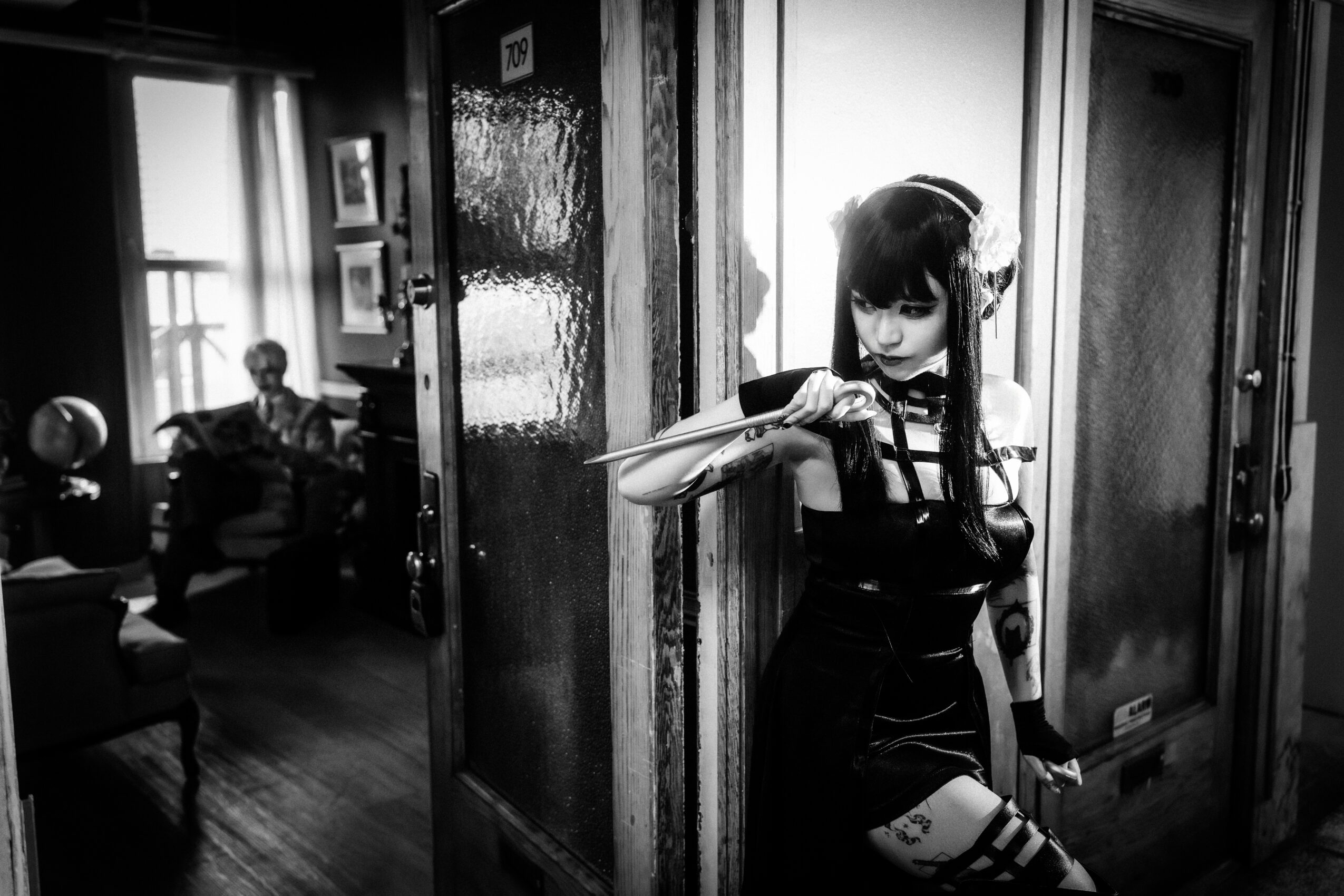 A girl with long dark hair and a bow, standing in a hallway, holding a violin, with people sitting in a room behind her.