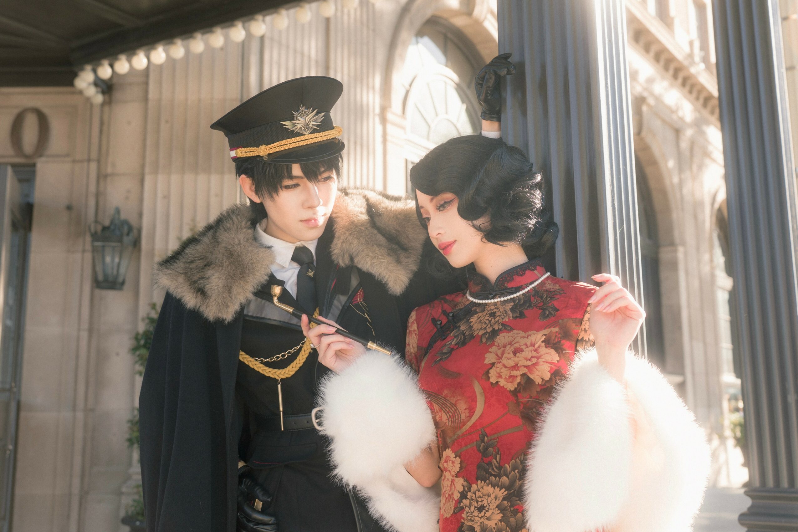 Two people in vintage military and traditional attire stand outdoors near columns and a building, looking at a phone.