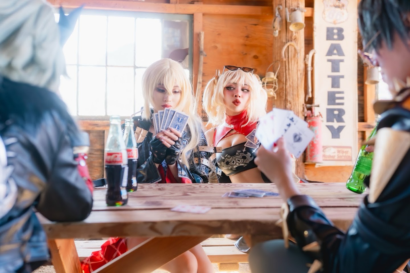 Two women with blonde wigs and costumes sit at a wooden bar, holding playing cards, with bottles on the counter and a window behind them.