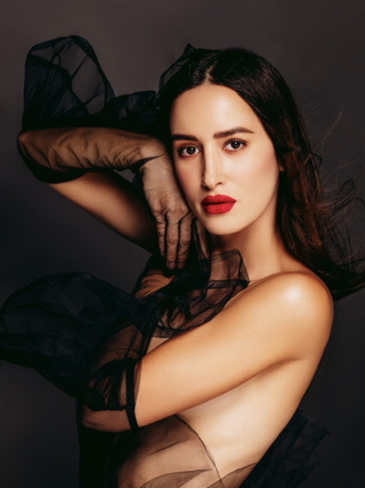 Woman with dark hair and red lipstick poses with her arm across her face, wearing sheer black fabric, against a dark background.
