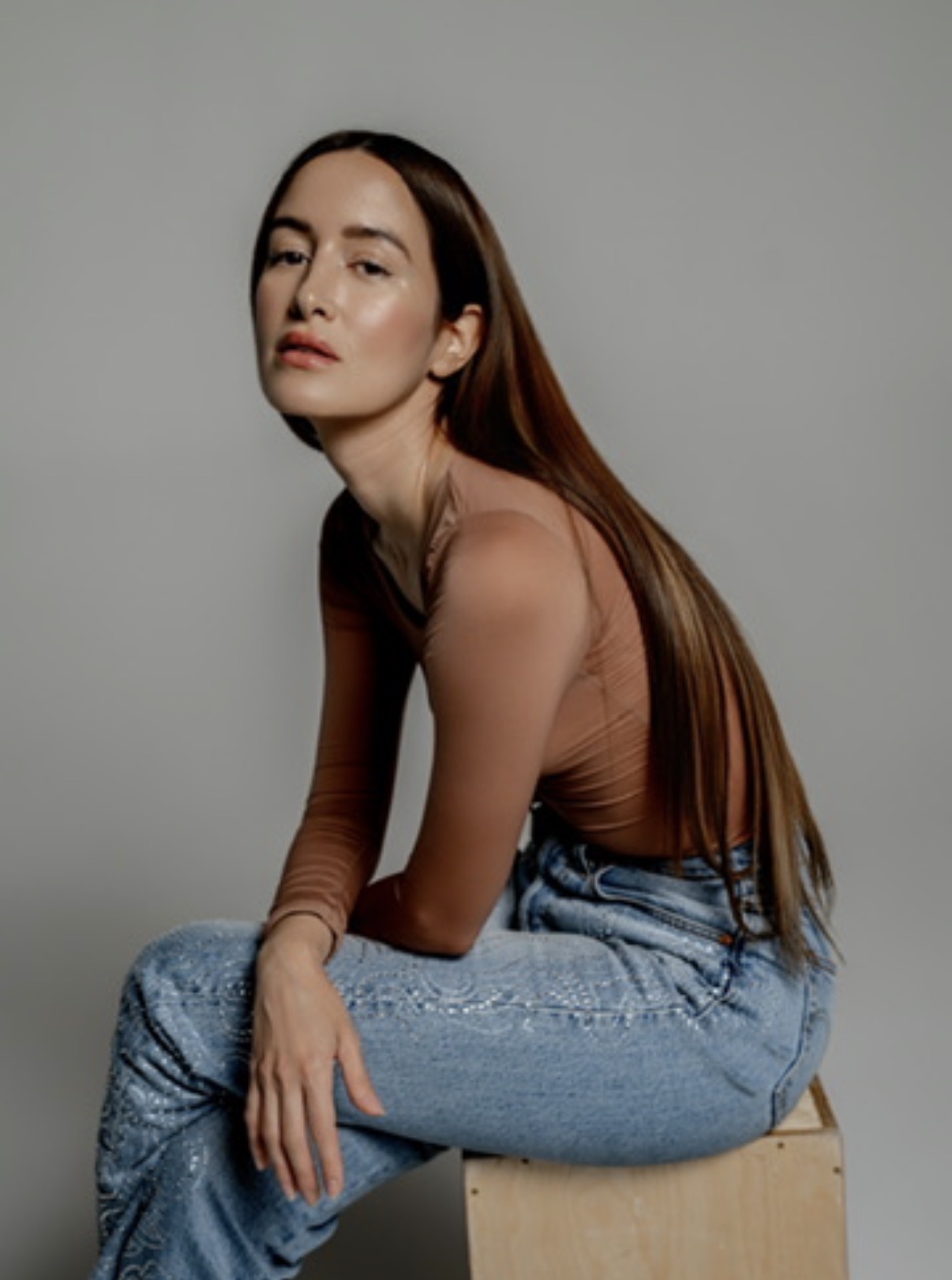 Young woman with long brown hair sitting on a wooden box, wearing a brown top and blue jeans, against a plain background.