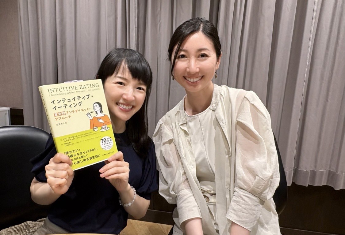 Two women smiling, one holding a yellow book, sitting in front of a curtain backdrop.