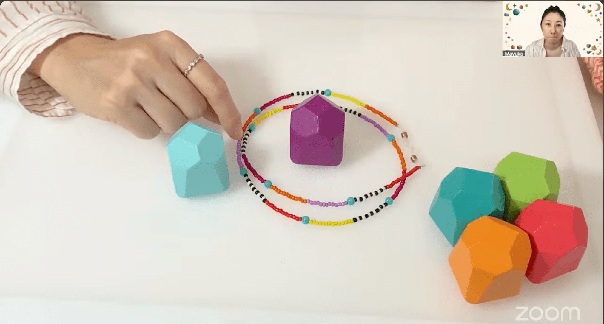 Person's hand with rings points to colorful geometric shapes on a white surface, with a beaded circle and a small video inset.