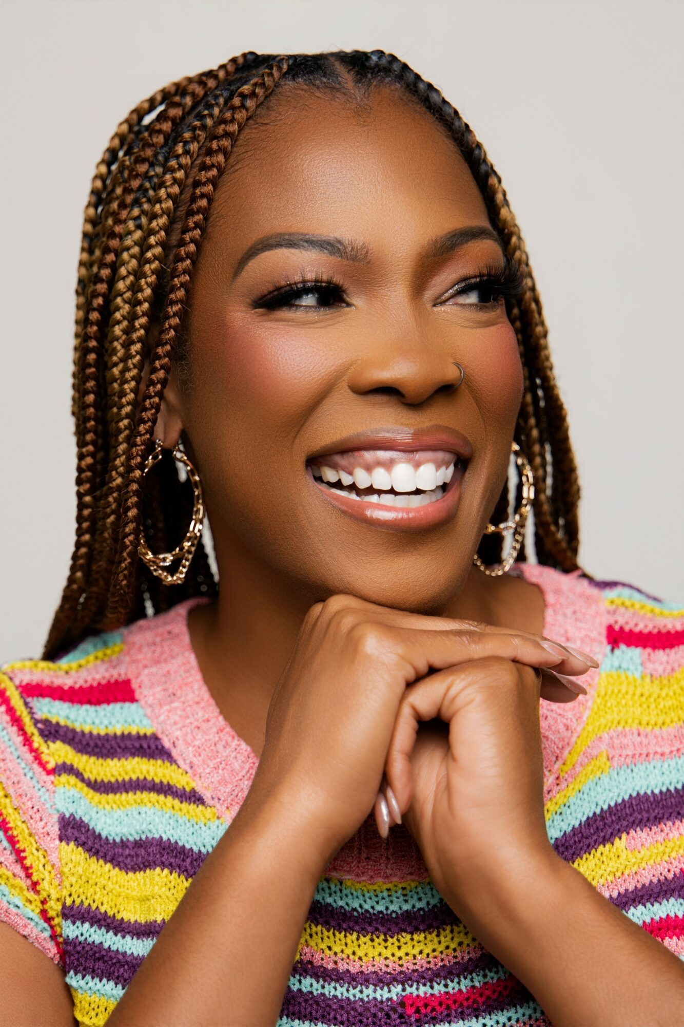 Smiling woman with braided hair, wearing colorful striped sweater, resting chin on hand, looking to the side.