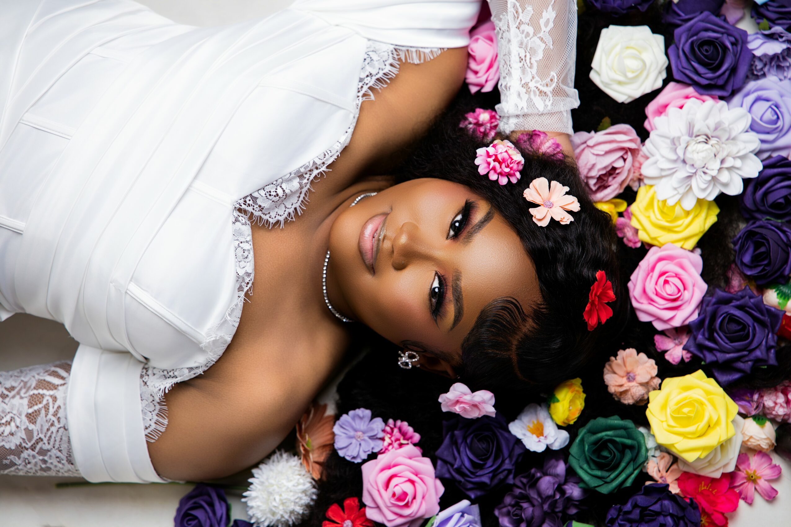 Woman lying on colorful flowers, wearing a white dress with lace details, looking at the camera.
