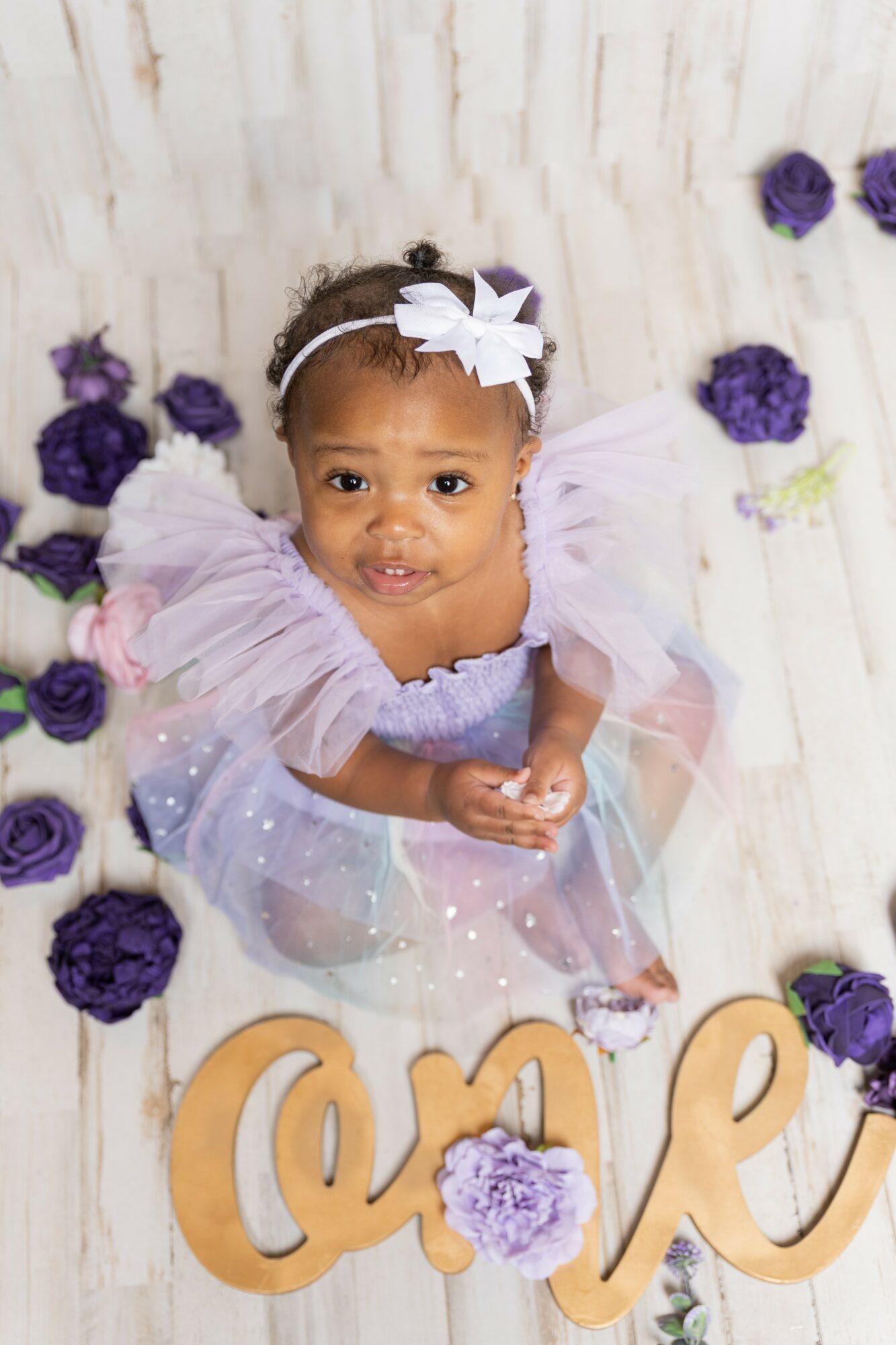 Young girl in a colorful dress with a headband, surrounded by purple flowers, looking up at the camera.