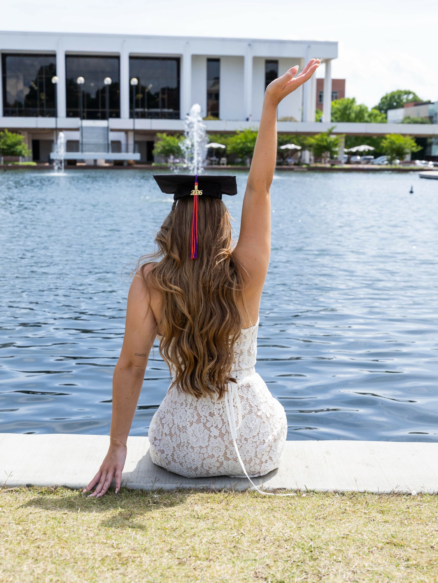 Woman with long hair sitting by a lake, raising one arm, with a modern building and fountain in background.