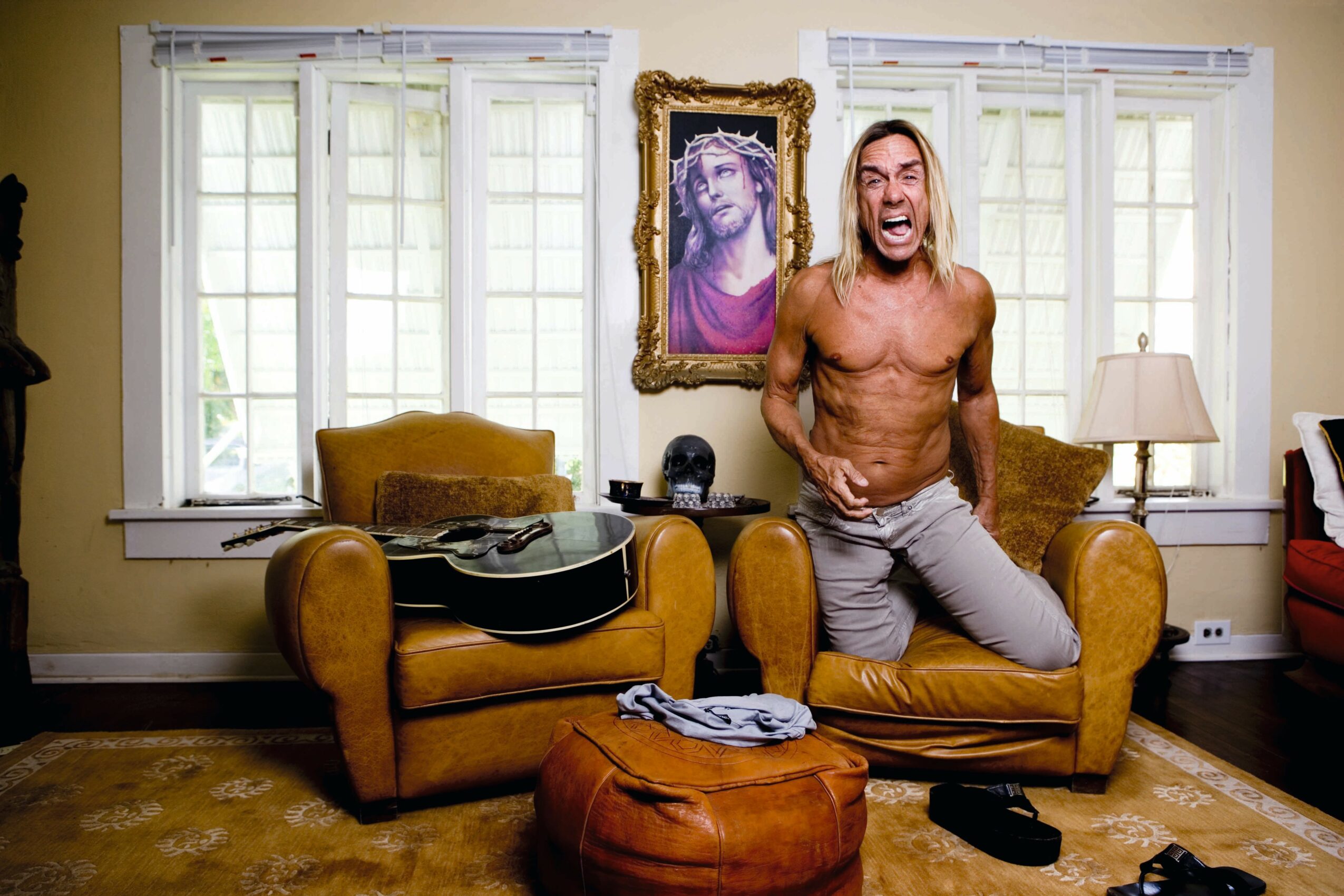 A shirtless man with long hair sits on a yellow sofa in a living room, smiling. There are windows, a guitar, and a painting behind him.