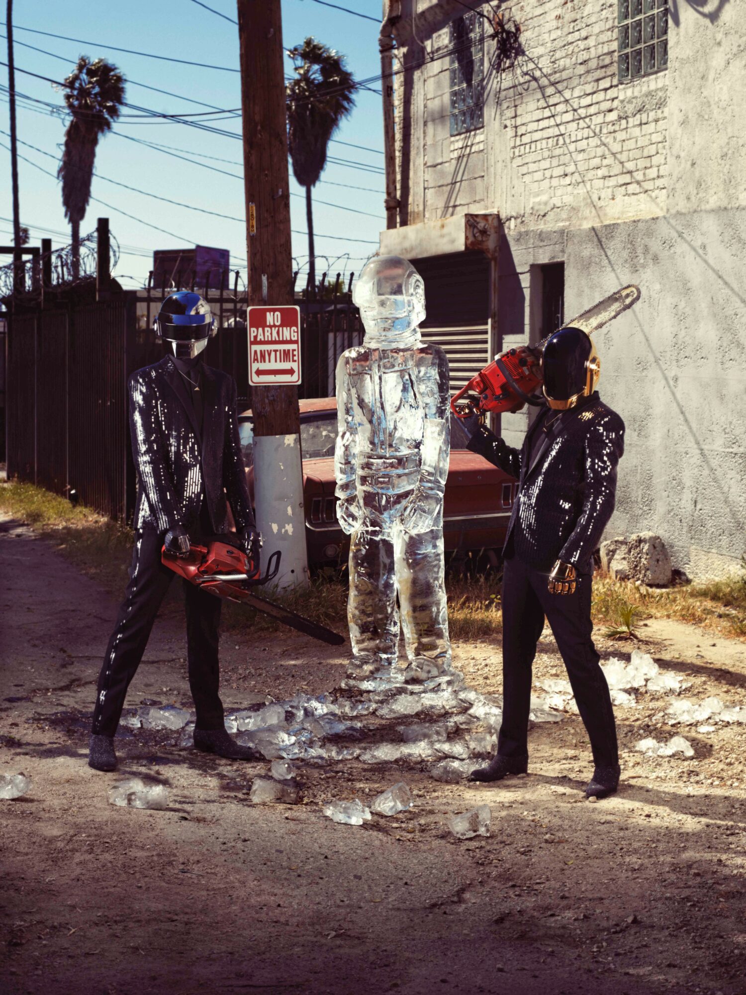 Two people in black outfits and masks stand beside a large ice sculpture of a human figure on a street.