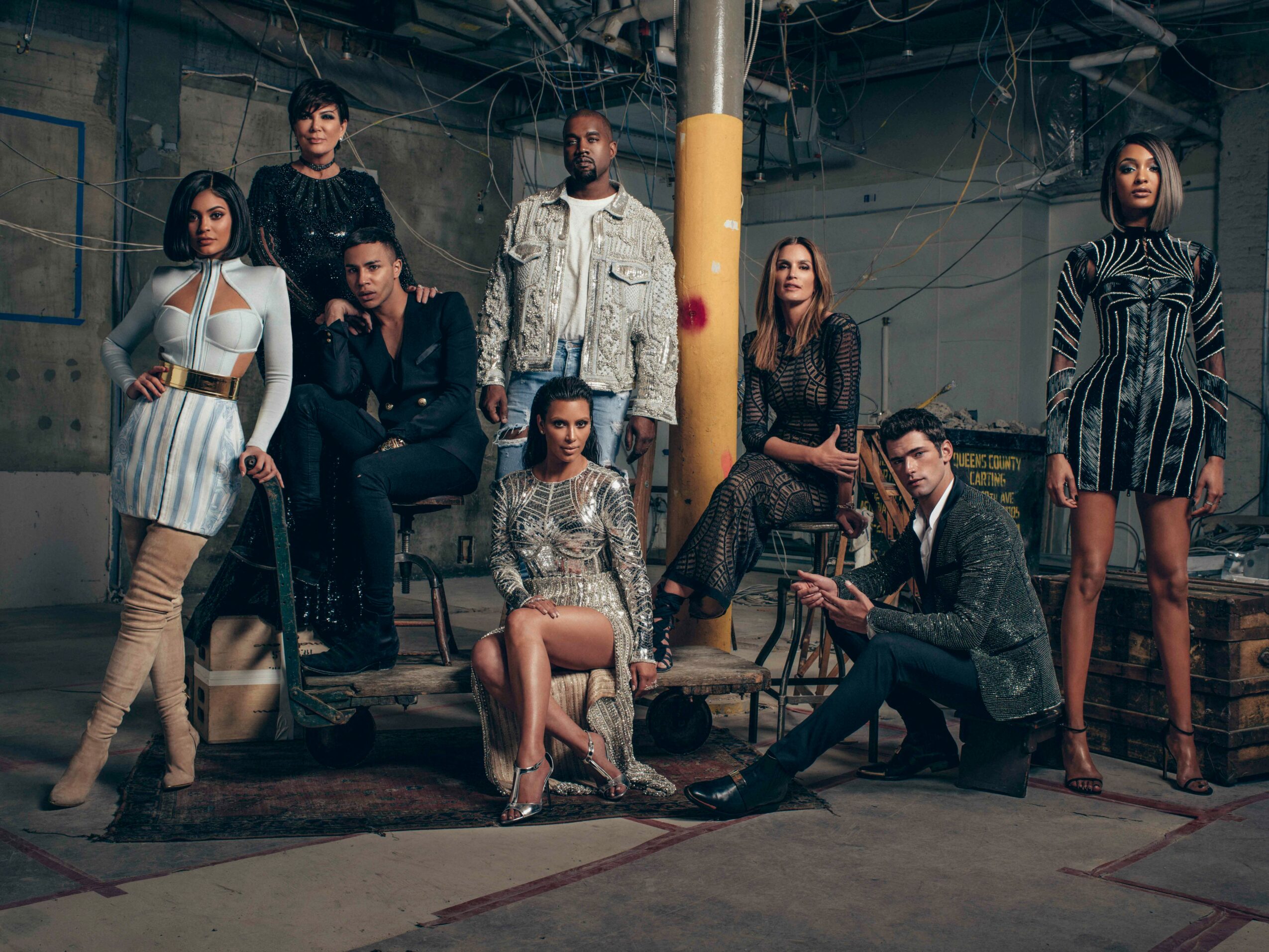 Group of nine diverse people in stylish clothing pose in an industrial setting with exposed pipes and a yellow pillar.