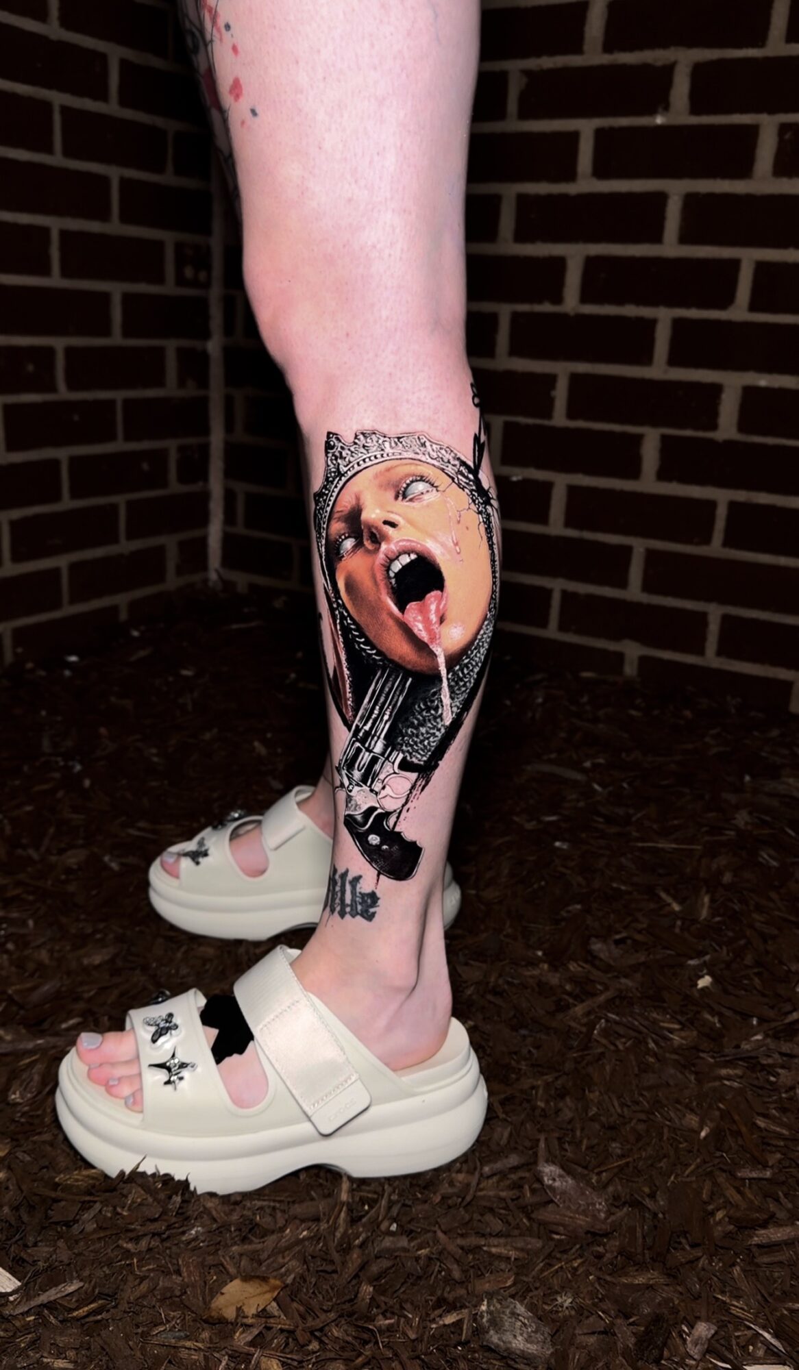 Tattoo of a woman's face with mouth open and tongue out on person's lower leg, wearing white sandals, brick wall background.