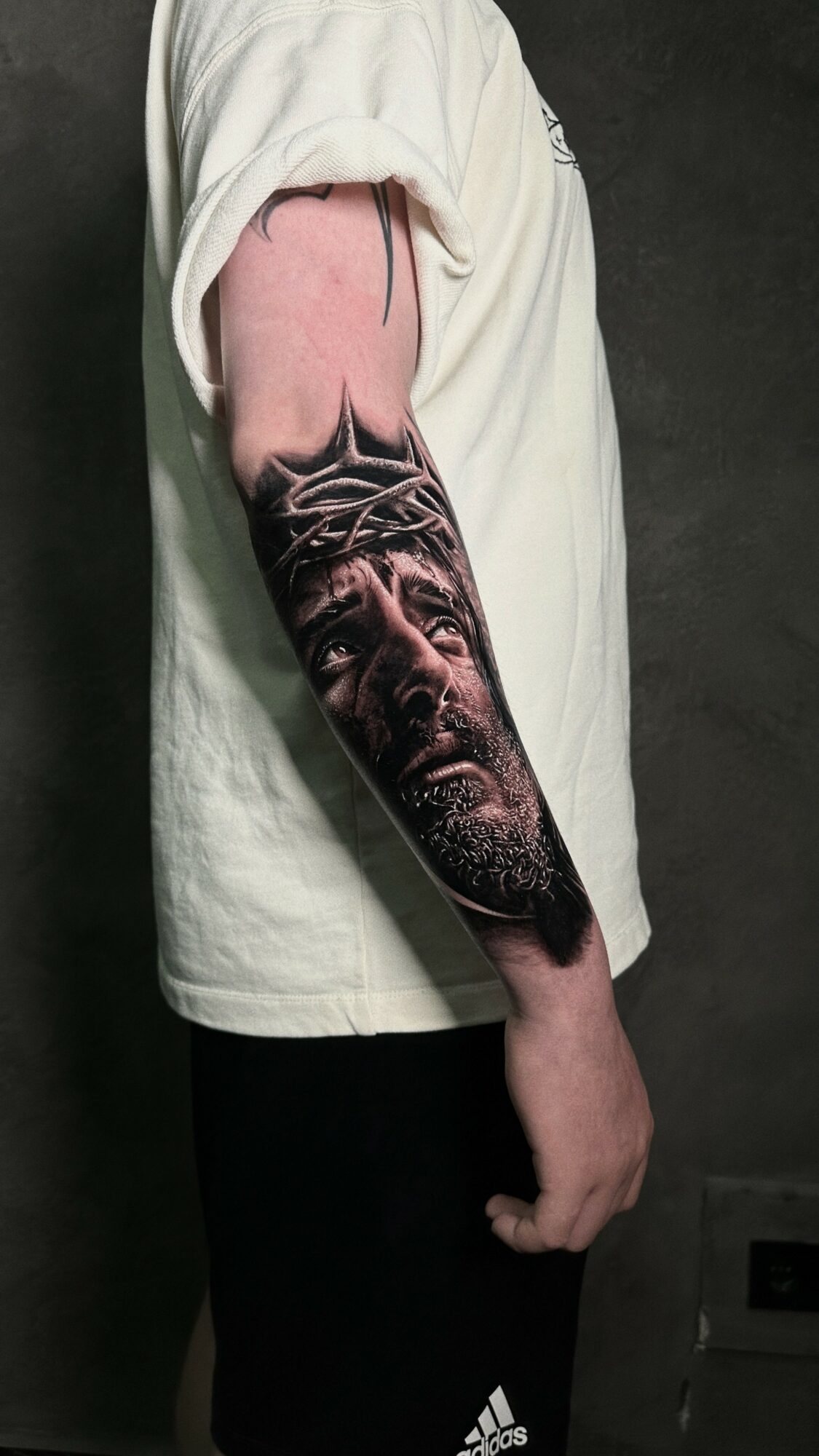 Person with a detailed black and gray tattoo of a face on their forearm, wearing a white shirt and black pants.