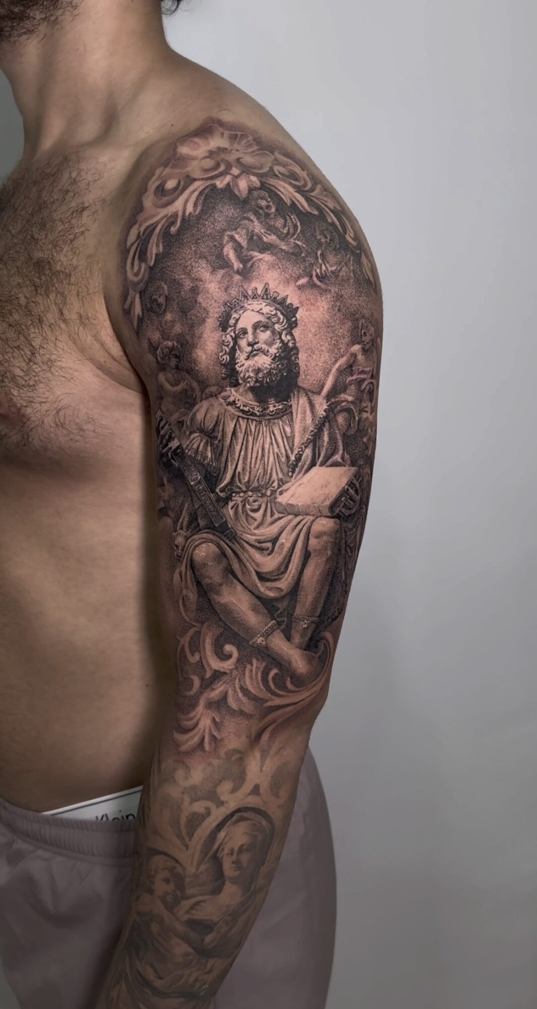 Tattoo of a bearded man with a halo, seated and holding a book, on a person's upper arm and shoulder.