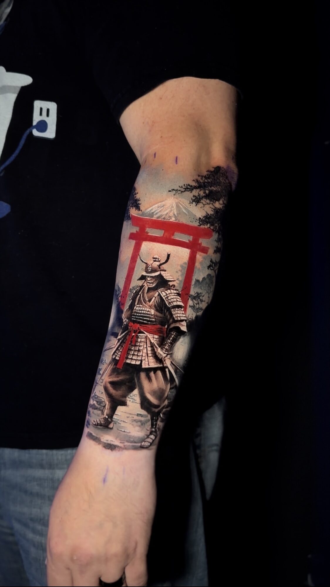 Tattoo of a samurai warrior in armor on forearm, with a red torii gate and landscape background.
