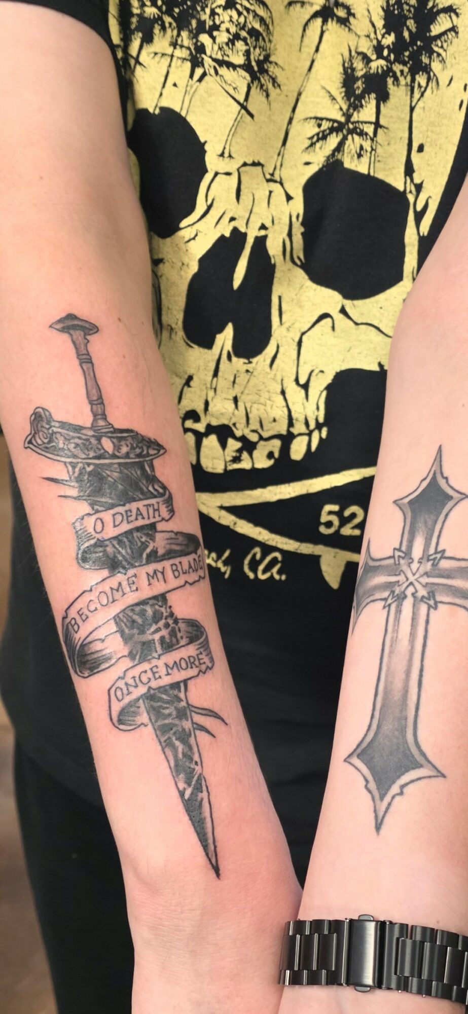 Two arms with tattoos: one with a sword and banner, the other with a stylized dagger, against a skull shirt background.