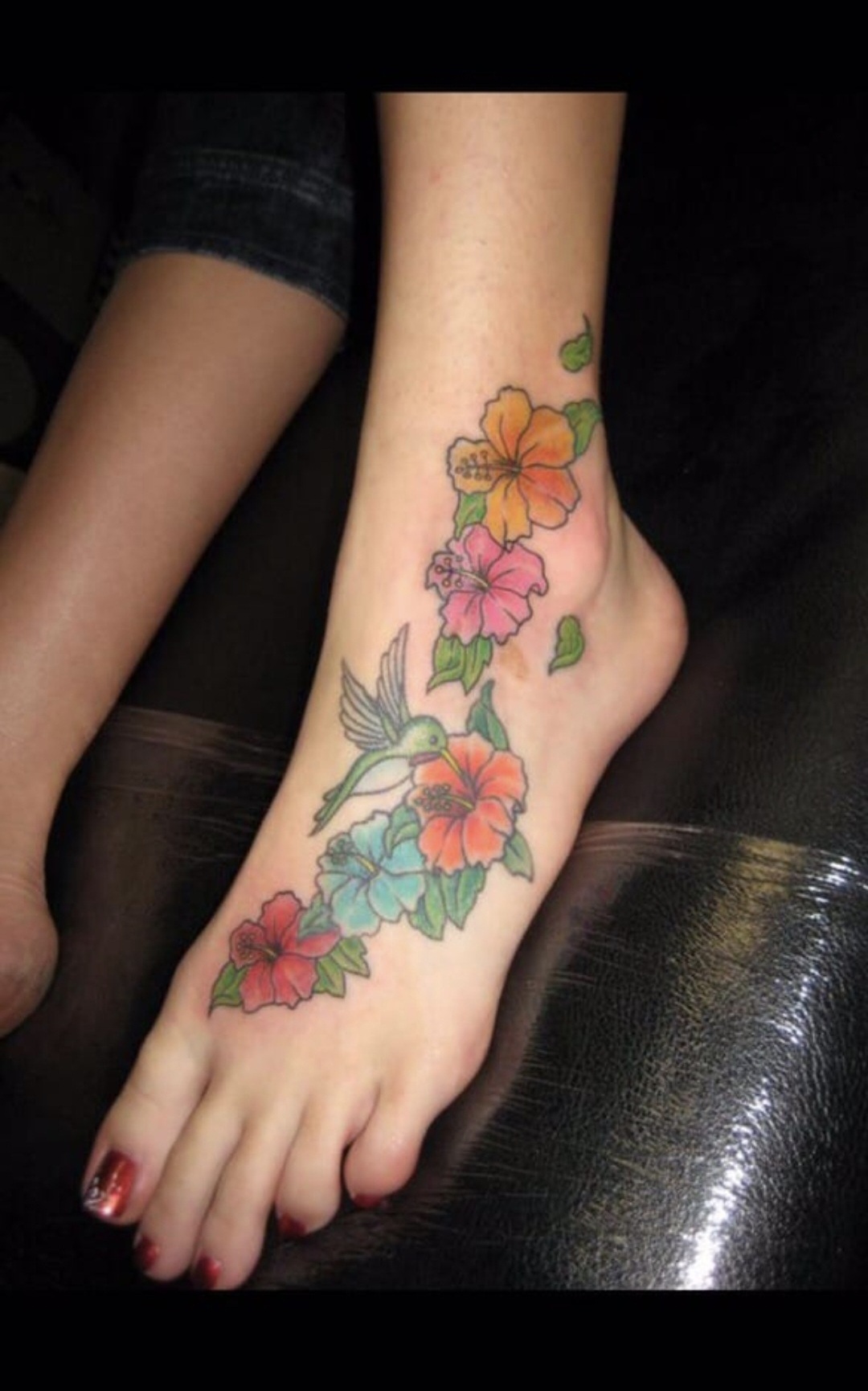 Foot with colorful floral tattoo, including pink, orange, and yellow flowers, and green leaves, resting on a black surface.