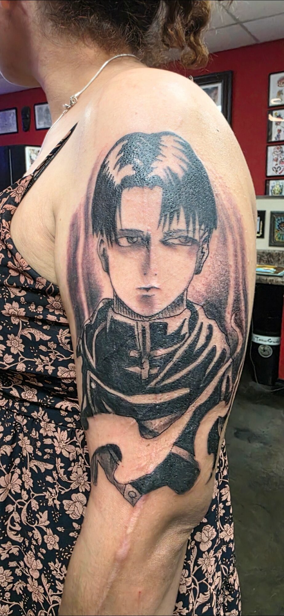 Tattoo of a character with dark hair and intense expression on upper arm, with a dark background and clothing, in a tattoo shop.