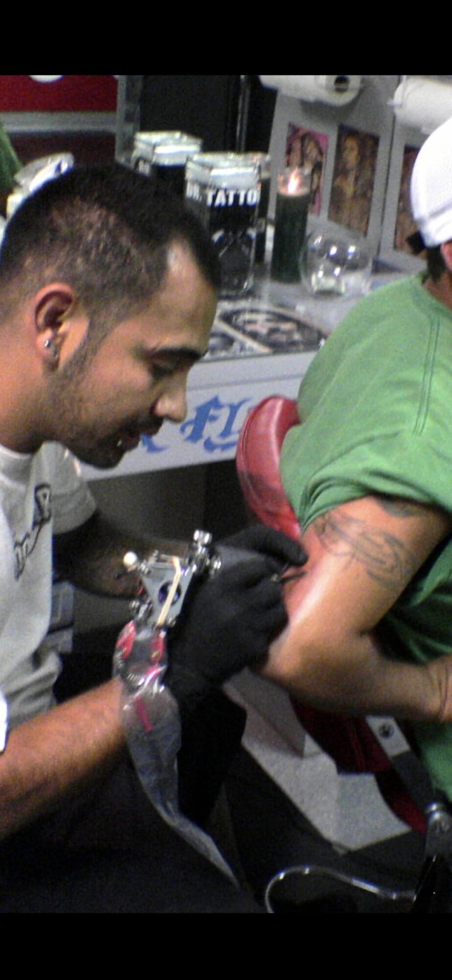 Tattoo artist tattooing a person's arm in a busy shop, with tattoo supplies visible in the background.