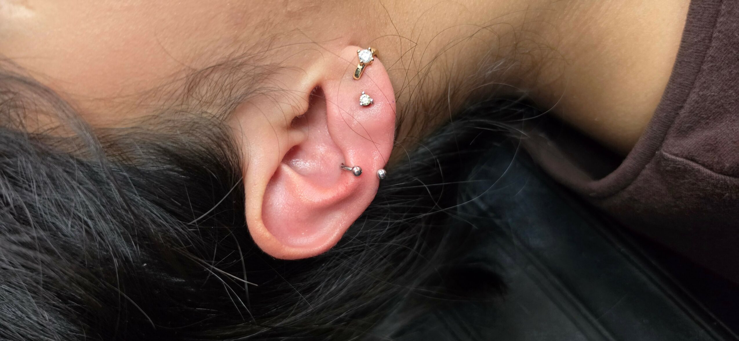 Close-up of a person's ear with multiple piercings, including a helix and lobe piercings, with dark hair nearby.