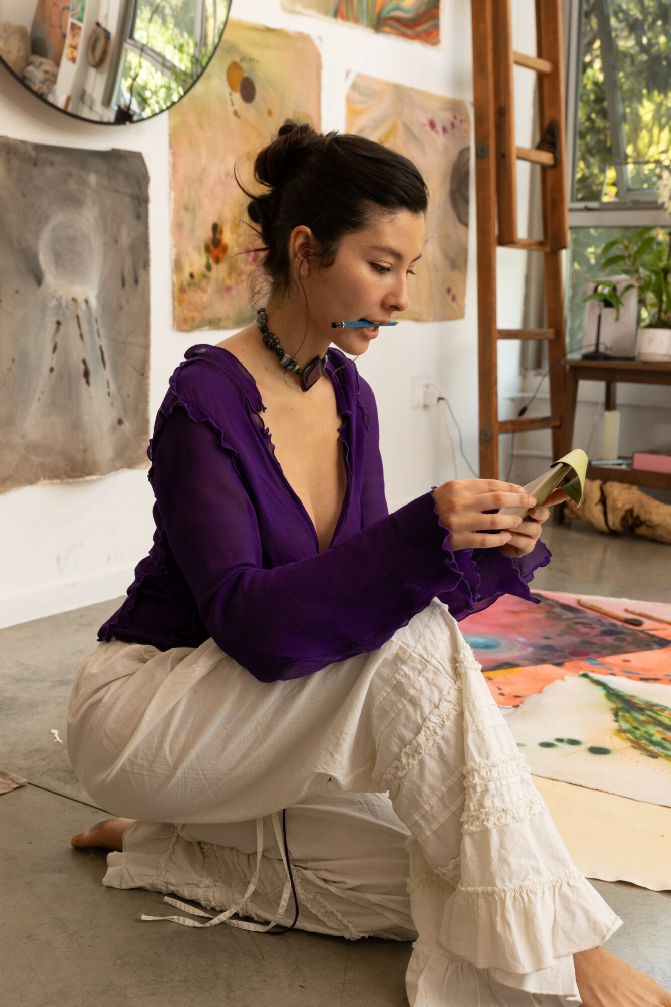 Woman sitting on floor, painting with watercolor, surrounded by artwork and art supplies, in a bright studio with plants and window.