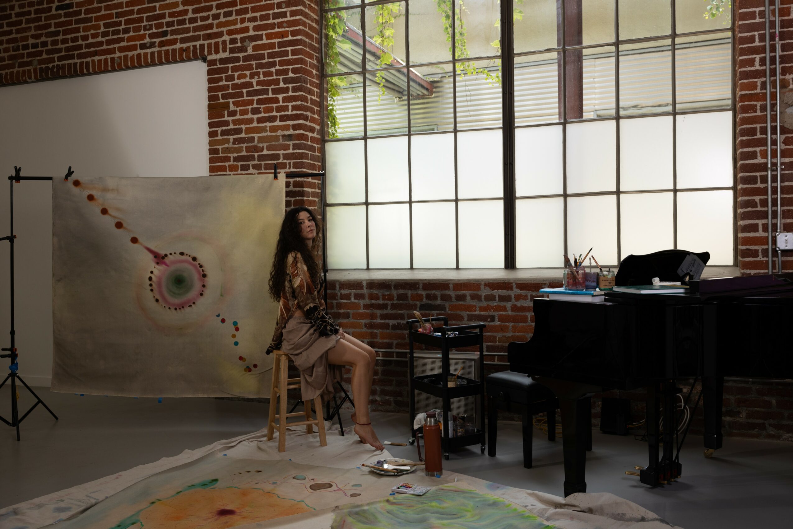 Woman sitting on a stool in a room with large windows, brick walls, and a black piano, with a backdrop and photography equipment.