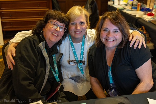 Three women sitting together, smiling, with arms around each other at a table in a room with wooden stairs and tables in the background.