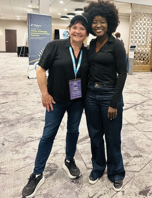 Two women standing together smiling in an indoor conference setting, one with short dark hair and the other with curly hair, both wearing black tops and jeans.