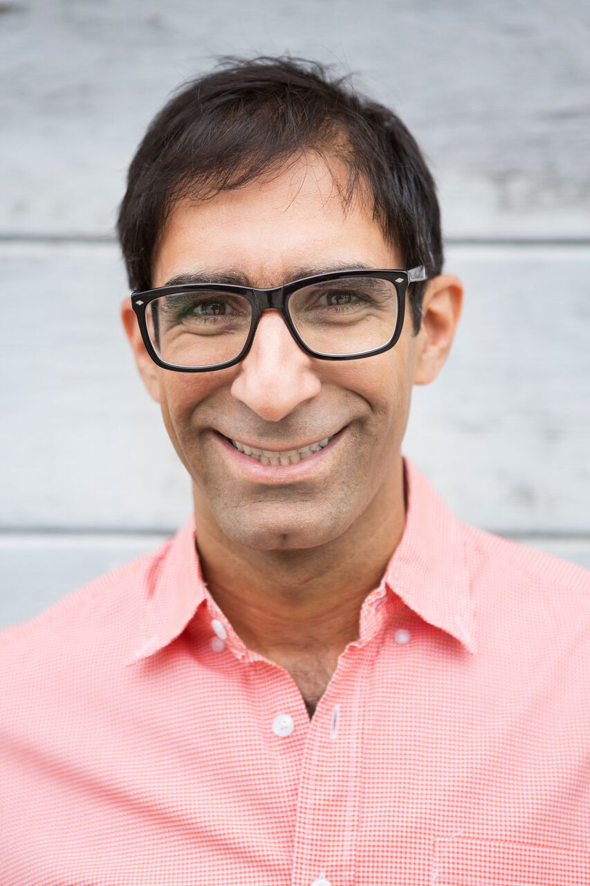 Man with glasses smiling, wearing a pink shirt, standing outdoors against a light background.