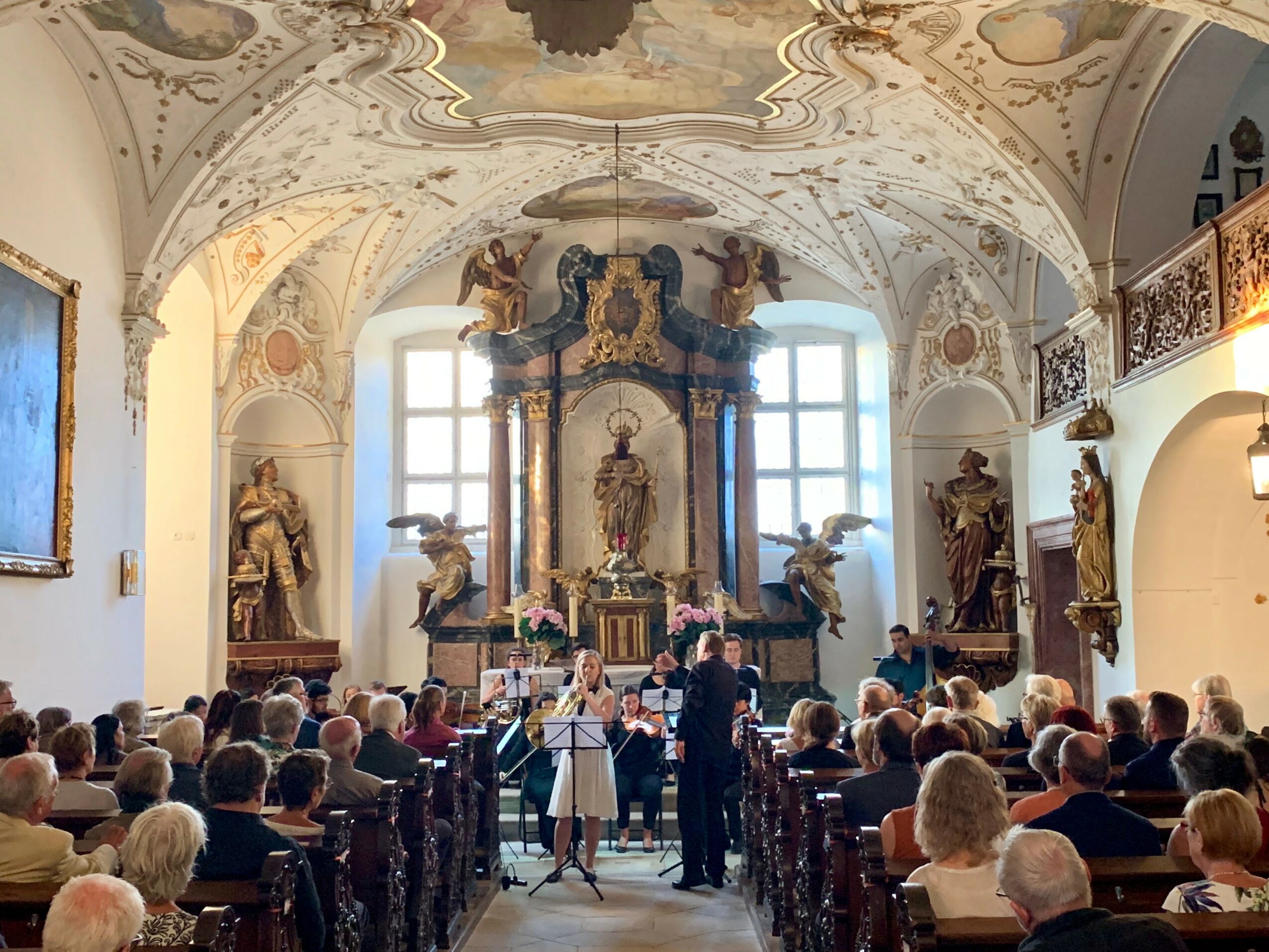 People attending a concert in a decorated church with statues and ornate ceiling, facing an altar with a statue and paintings.