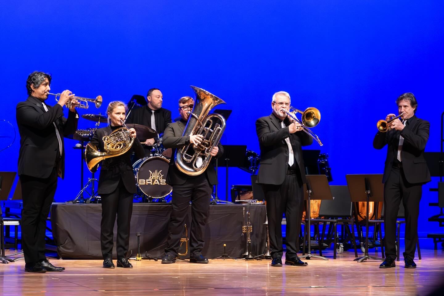 Six musicians playing brass instruments on stage with a blue background, some wearing suits, a drum set in the background.