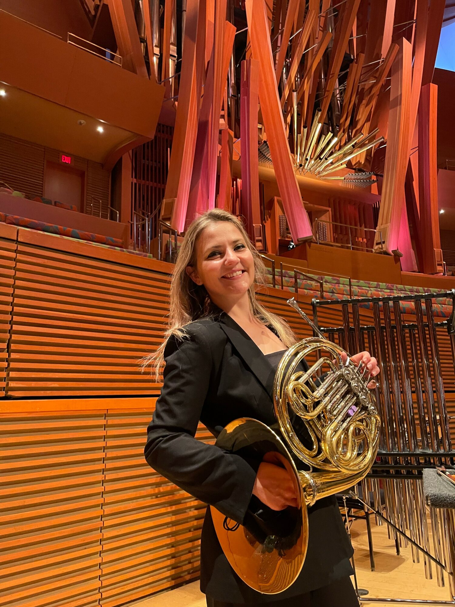 Woman holding a French horn and guitar, smiling, standing in a concert hall with orange and pink decorations.