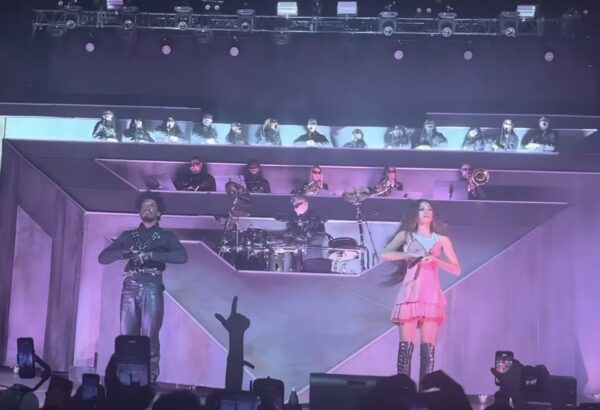 Stage with a singer and band, multiple musicians on elevated platform, audience in front, purple lighting, person making a peace sign.