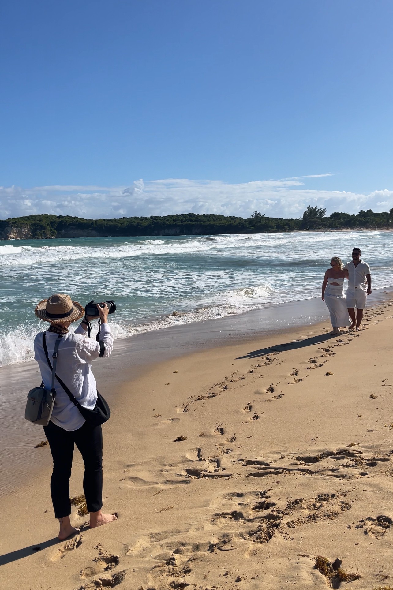 Person taking a photo of two people walking on the beach, ocean waves, blue sky, and distant shoreline.