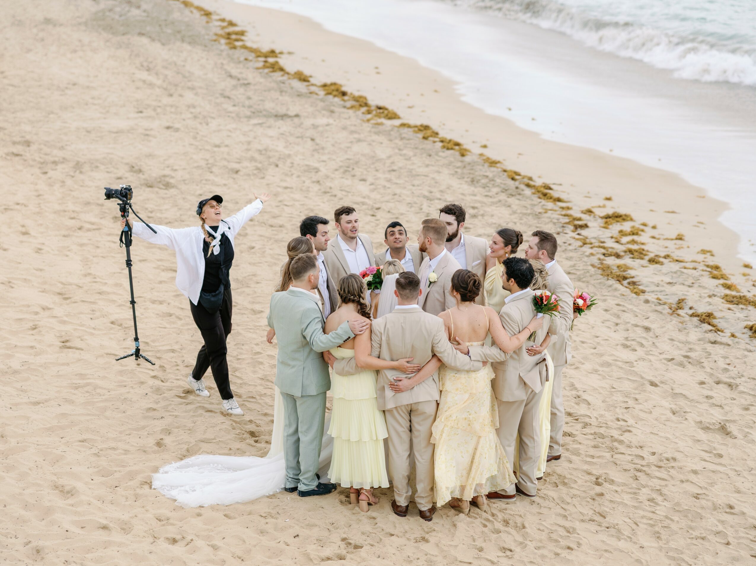 Group of people in wedding attire on sandy beach, with ocean waves in background, celebrating together.