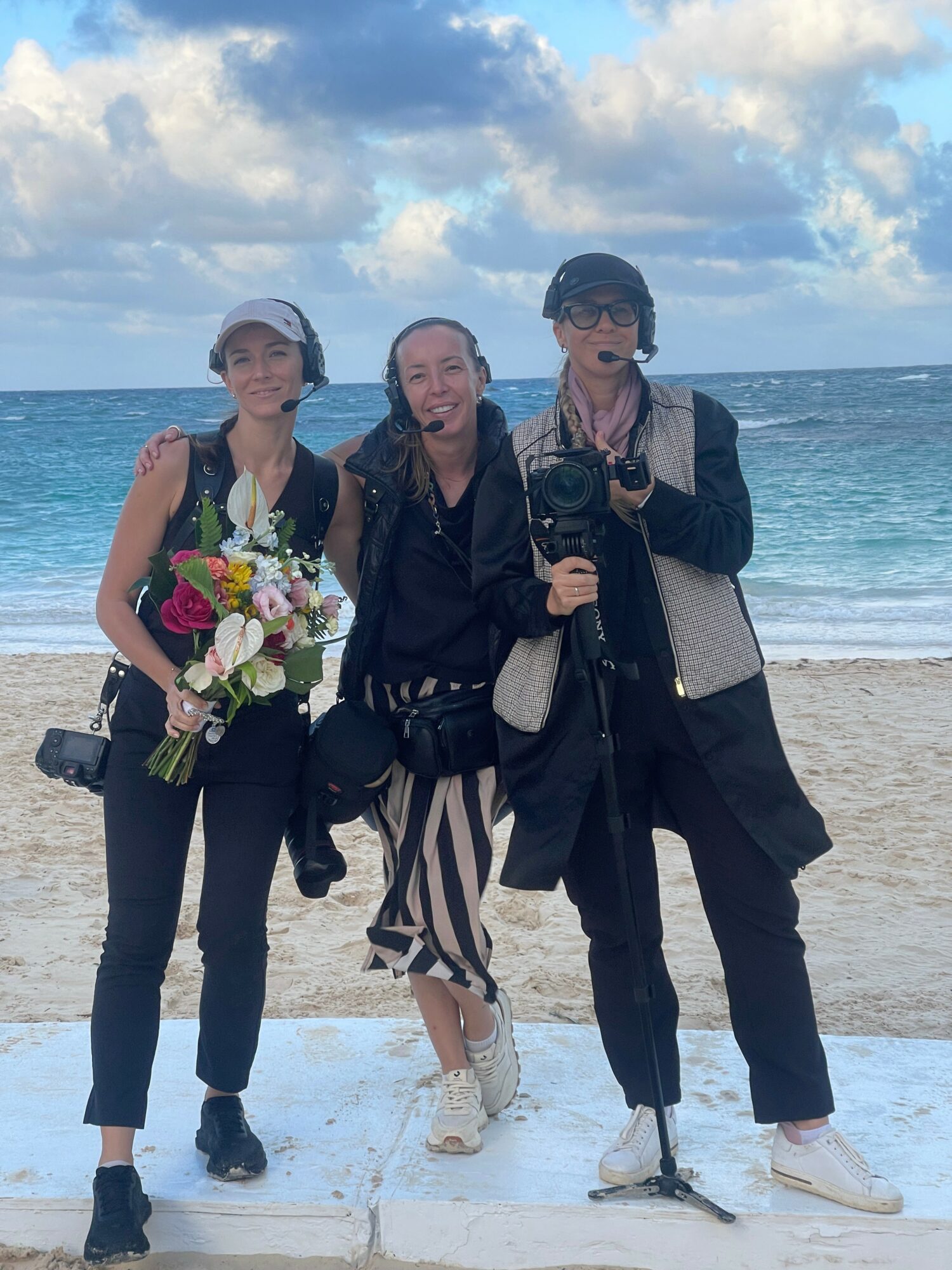 Three women standing on a beach with ocean and sky in the background, smiling, one holding a bouquet of flowers, one with a camera.