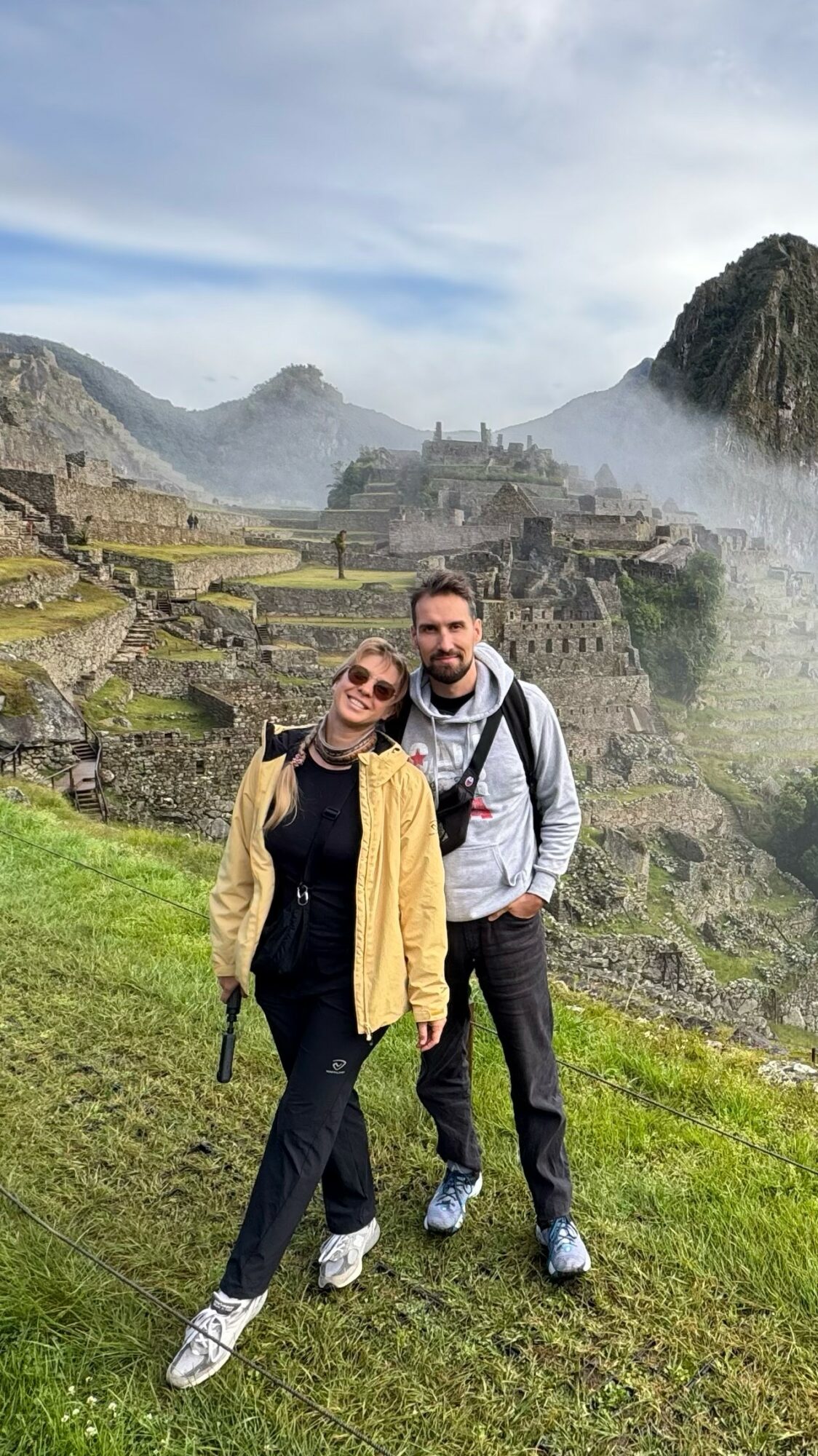 Two people standing on grass with ancient ruins and mountains in the background, cloudy sky above.