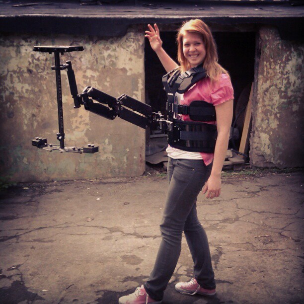 Young woman with long hair smiling, wearing a pink shirt and gray pants, standing outdoors with a camera rig.