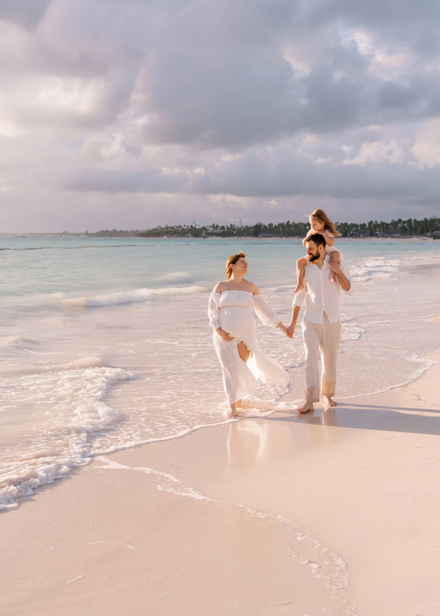 Family of four holding hands and playing on the beach with waves and cloudy sky in background.