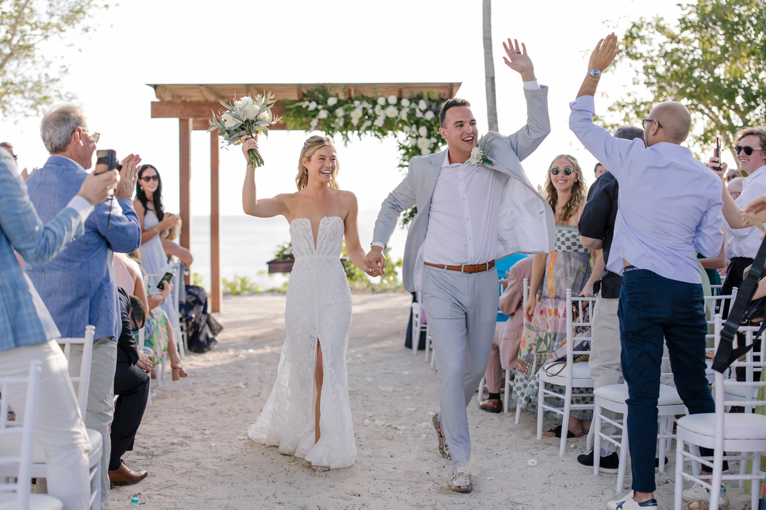 Bride and groom walk down aisle holding hands, guests cheer and celebrate outdoors, wedding arch decorated with flowers behind them.