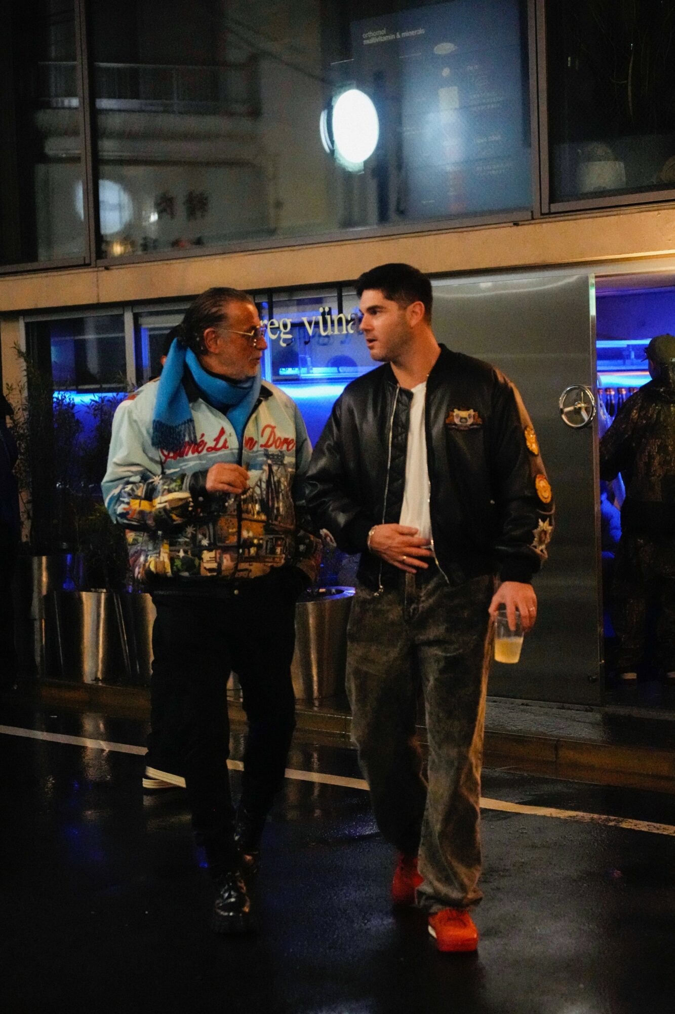 Two men stand and talk in a dimly lit indoor setting, one holding a drink, with a window and city lights in the background.