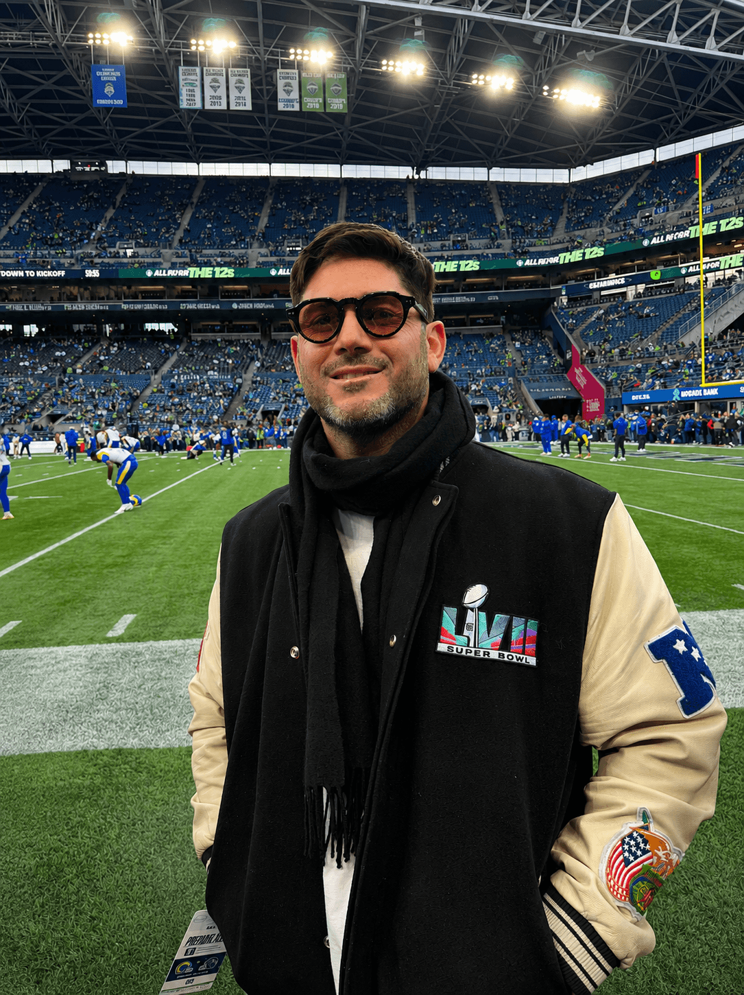 Man wearing sunglasses and a jacket stands on a football field with stadium and players in background.