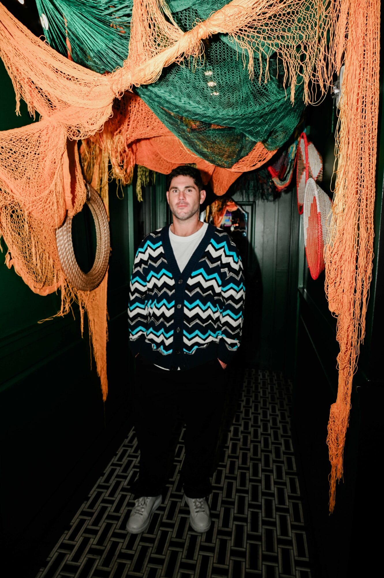 Young man standing in colorful fabric tunnel with hanging textiles and patterns, looking at camera.