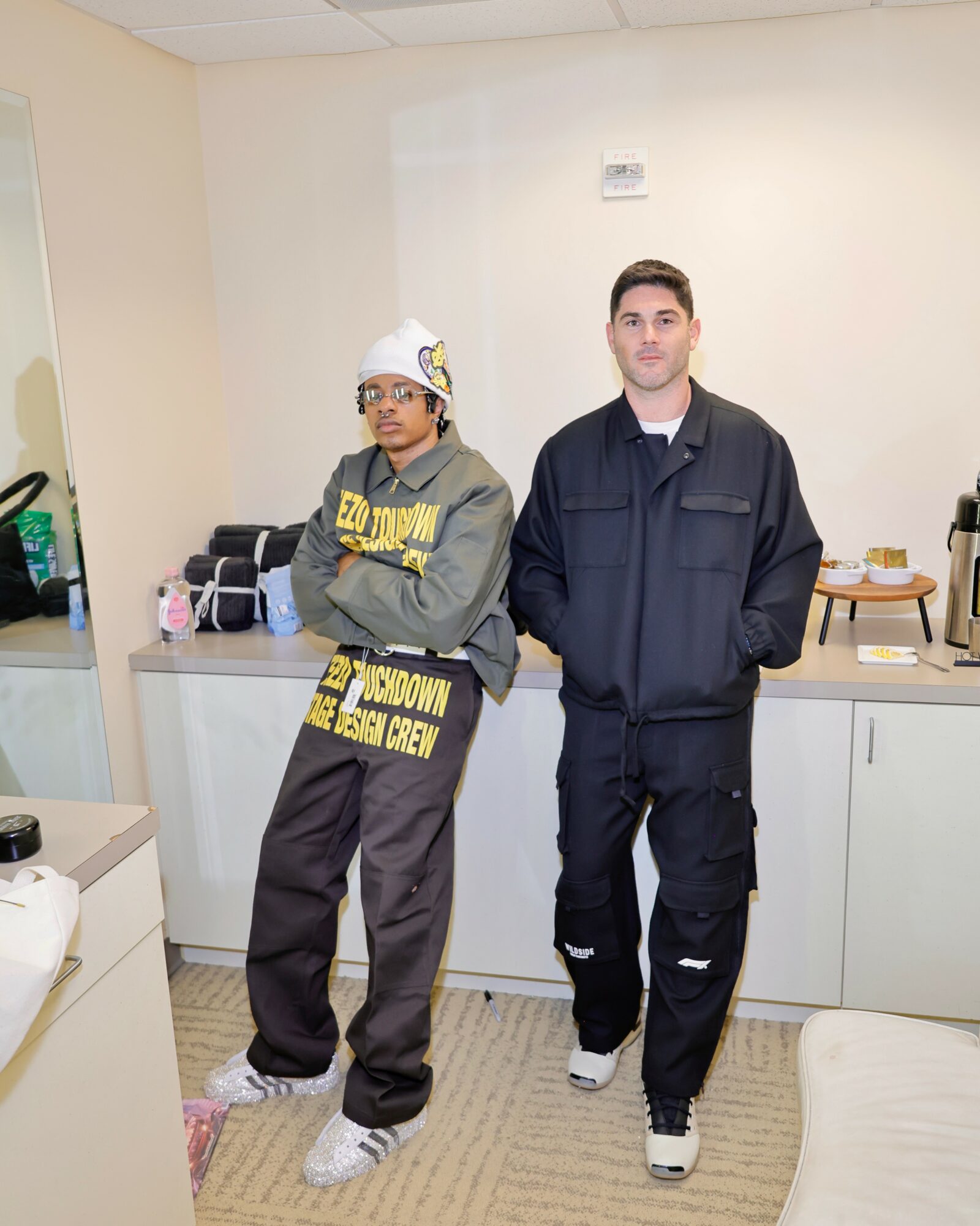 Two young men stand in a room with white walls and cabinets, one wearing a hoodie and glasses, the other in a jacket.