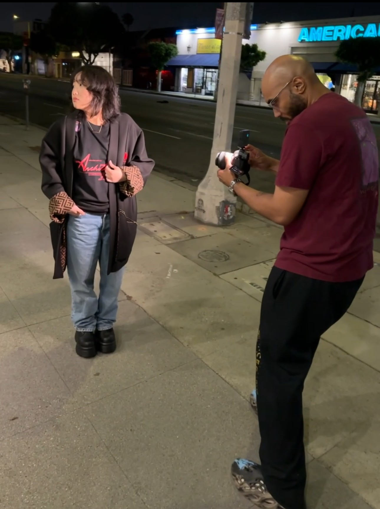 Two people standing on a sidewalk at night, one holding a camera, with a store and street in background.