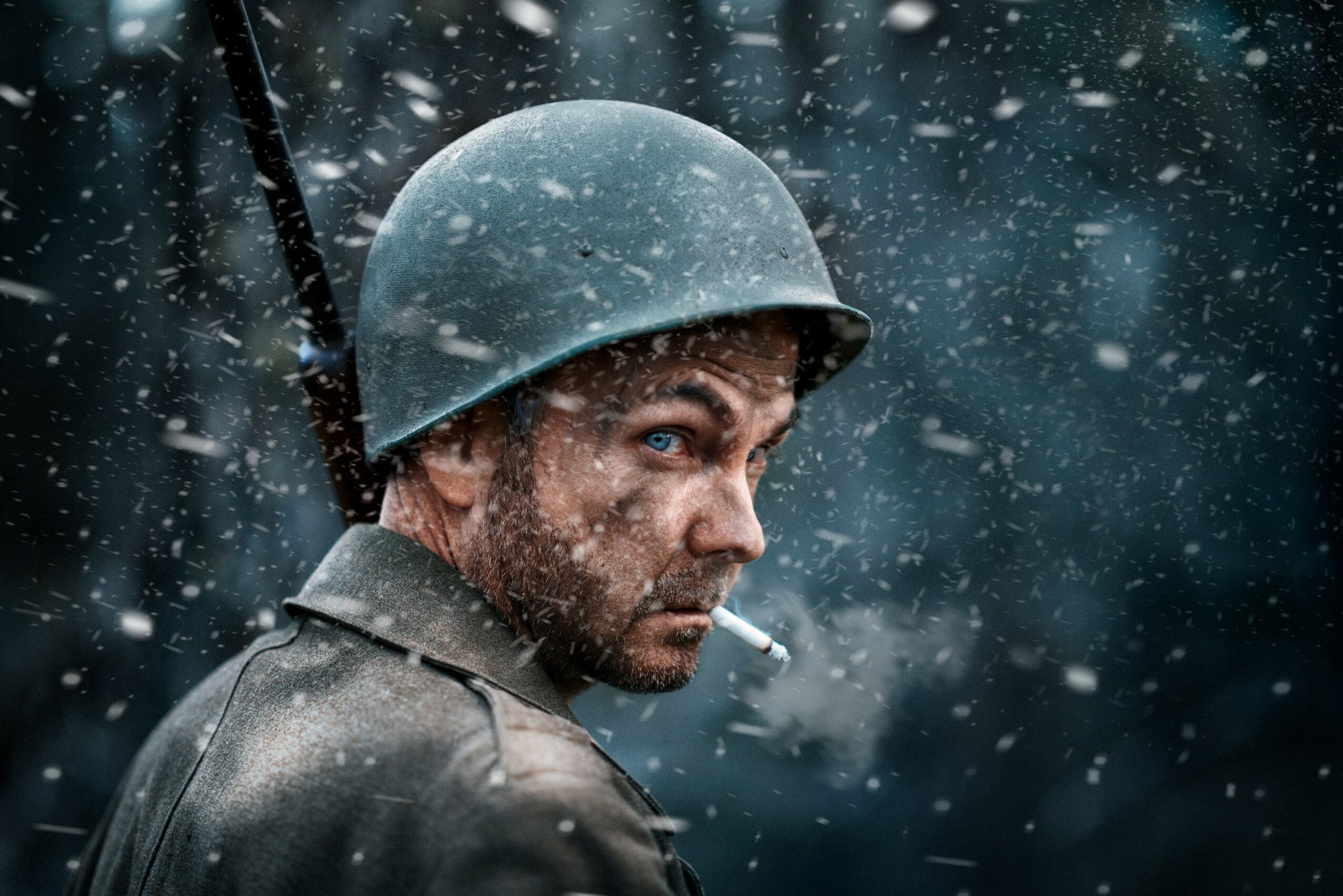 Man wearing a helmet with snow falling around him, holding a cigarette, with a serious expression.