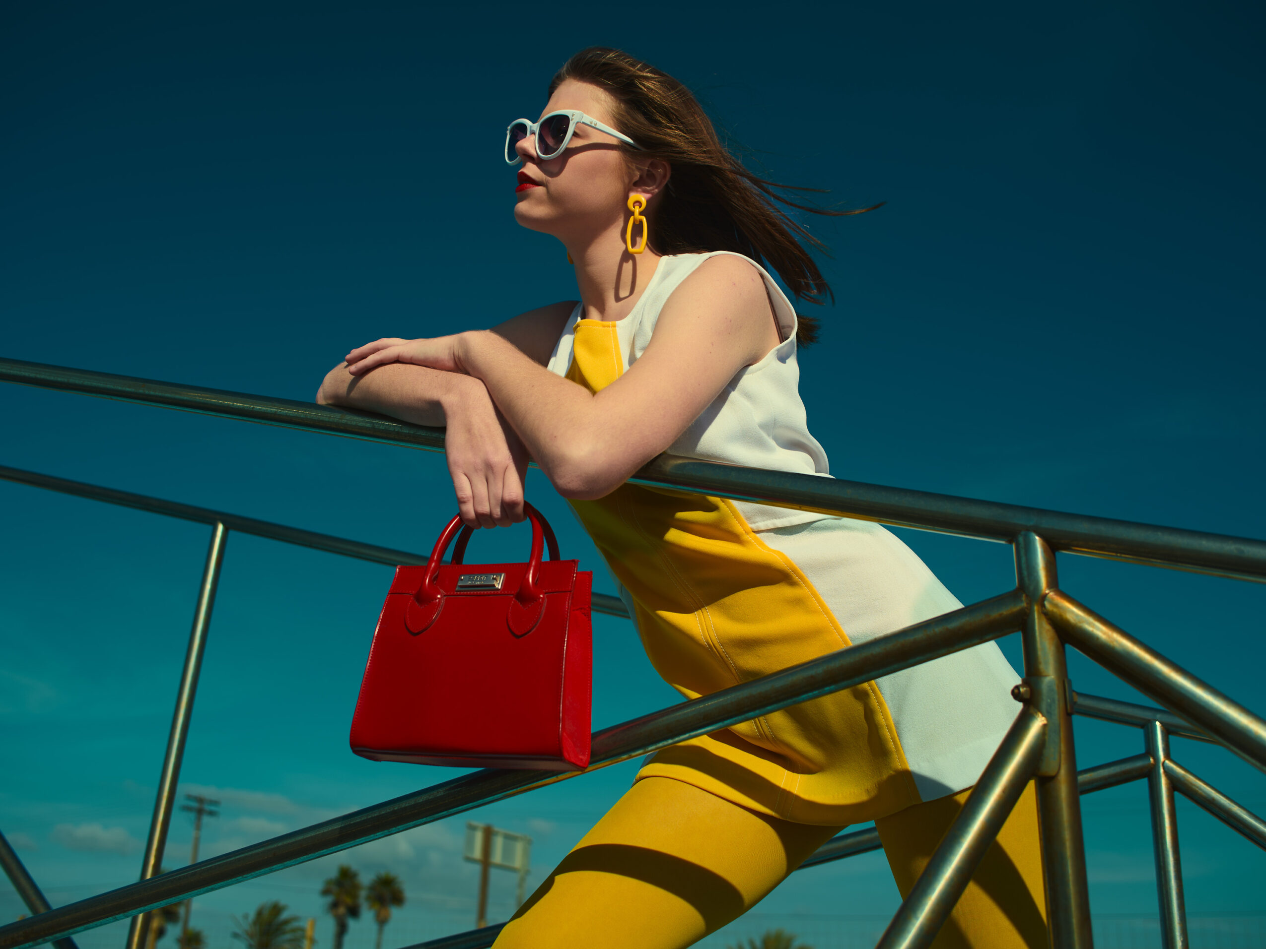 Woman with sunglasses and yellow dress leaning on a railing, holding a red handbag, against a blue sky.