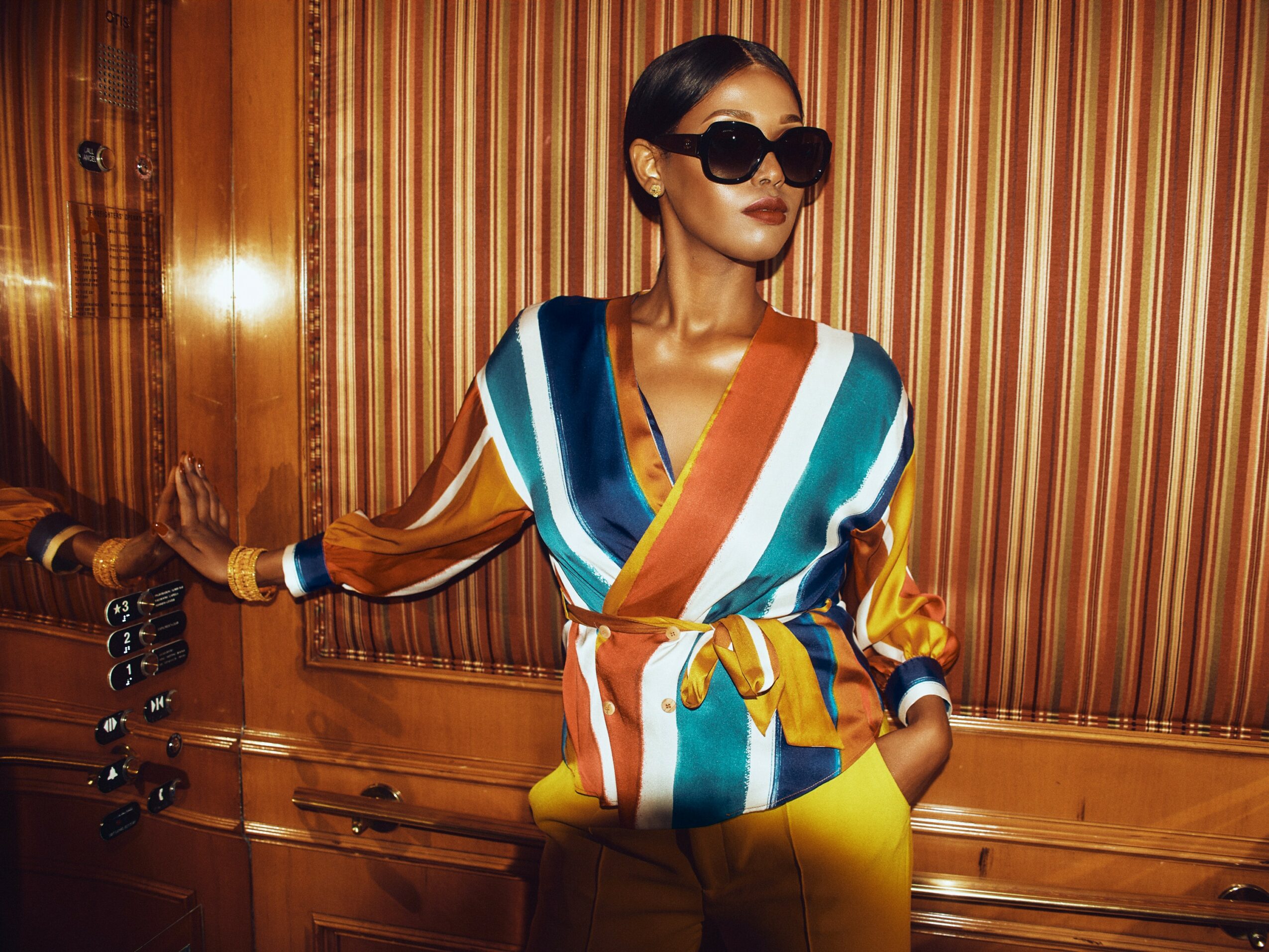Woman wearing sunglasses and a colorful striped blouse, standing against a wooden-paneled wall.